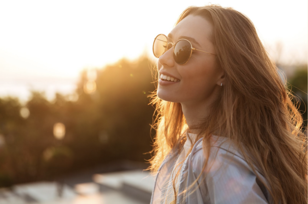 A young woman outside, smiling and wearing polarized sunglasses, as the sun shines.